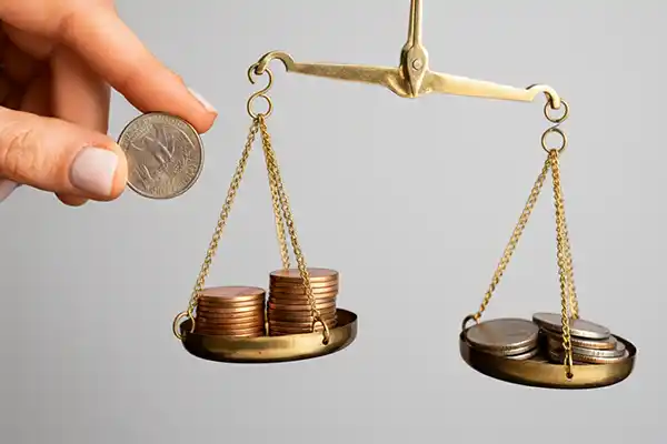 Mão adicionando moeda a uma balança de pratos equilibrada com pilhas de dinheiro, simbolizando a análise comparativa de regimes tributários feita pela Liz Consultoria em Campinas-SP.